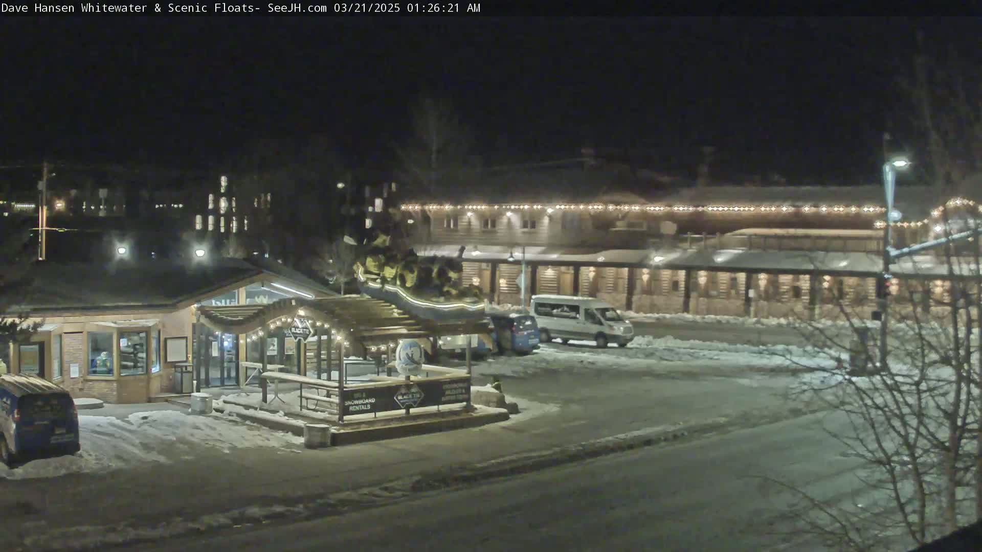 A nighttime view of a snow-covered parking lot with several vehicles, a small building with string lights, and a larger building in the background illuminated with lights.