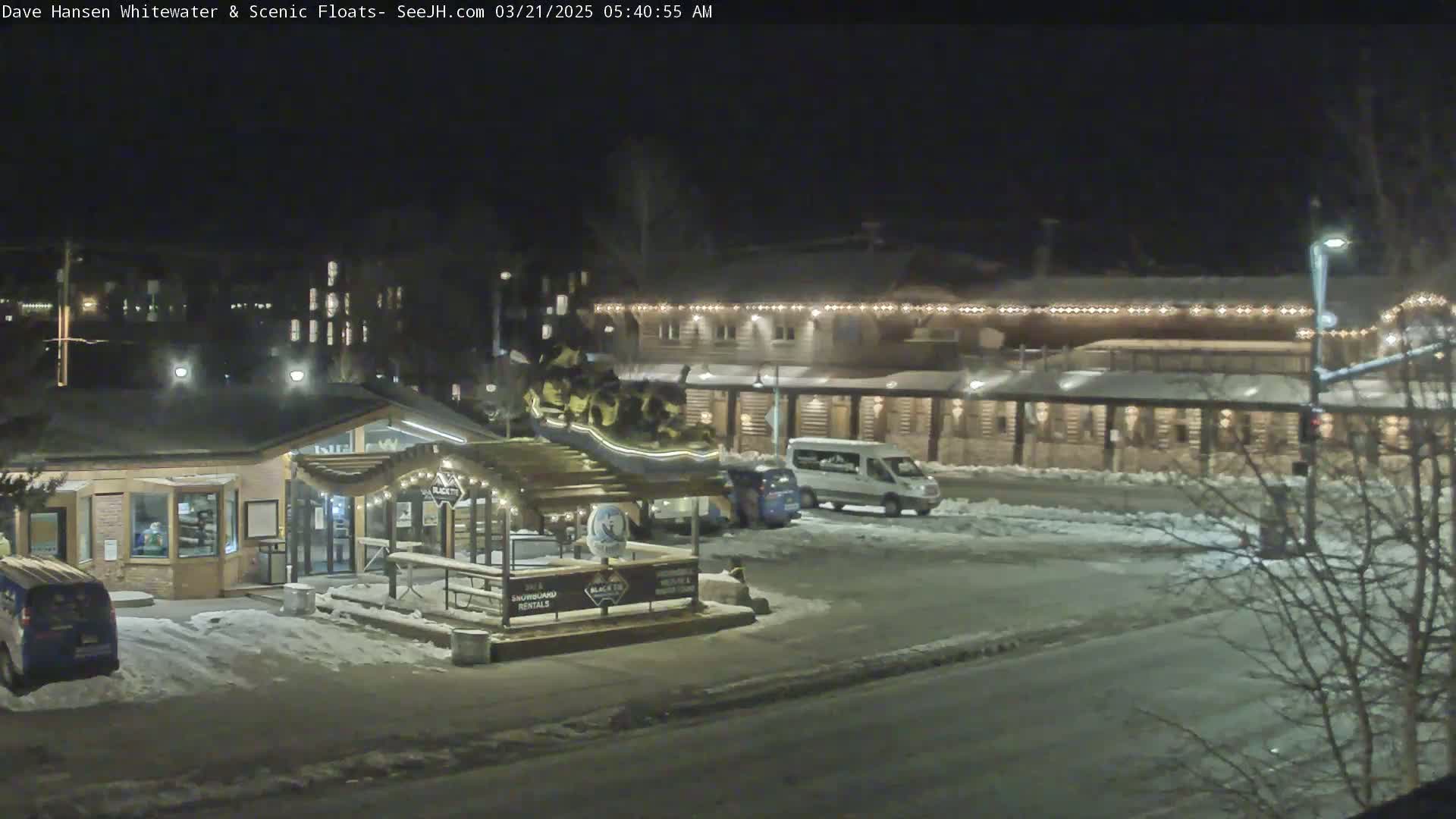 A nighttime scene of a snow-covered parking lot and street in front of illuminated buildings, with some vehicles parked.