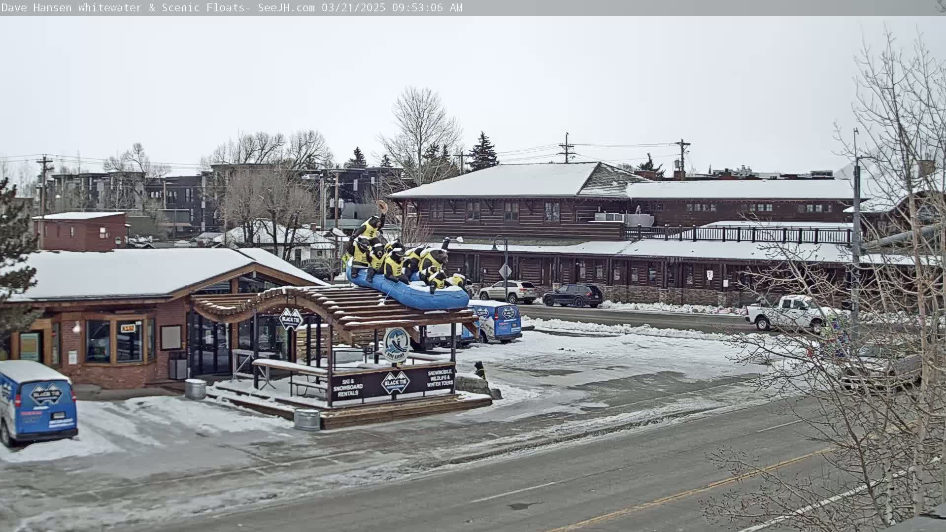A snow-covered street scene shows a building with a raft display on the roof, parked cars, and bare winter trees.