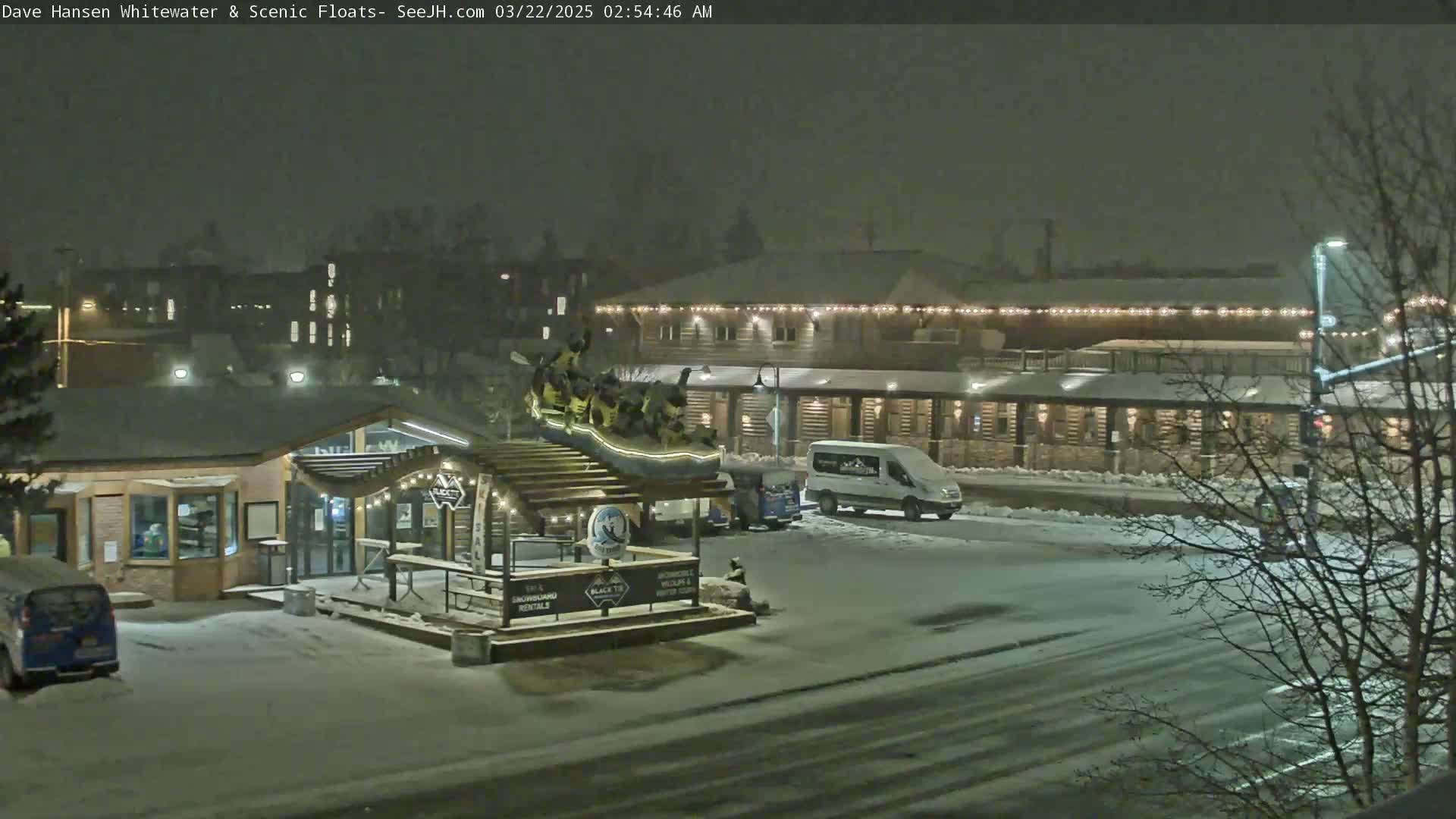 A snow-covered street scene at night shows a building with string lights, a small structure with a sign, and a van parked nearby.
