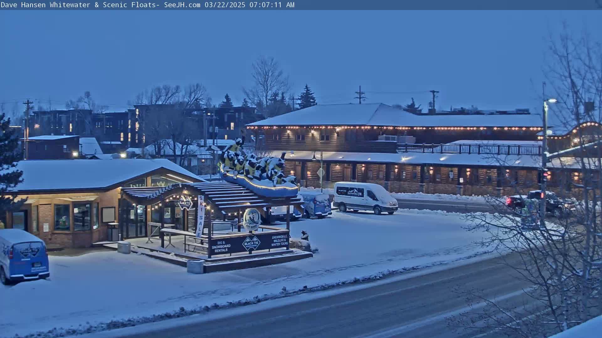 A snow-covered street scene at twilight shows a building with string lights, a ski and snowboard rental shop with a raft statue, and several vehicles parked nearby.