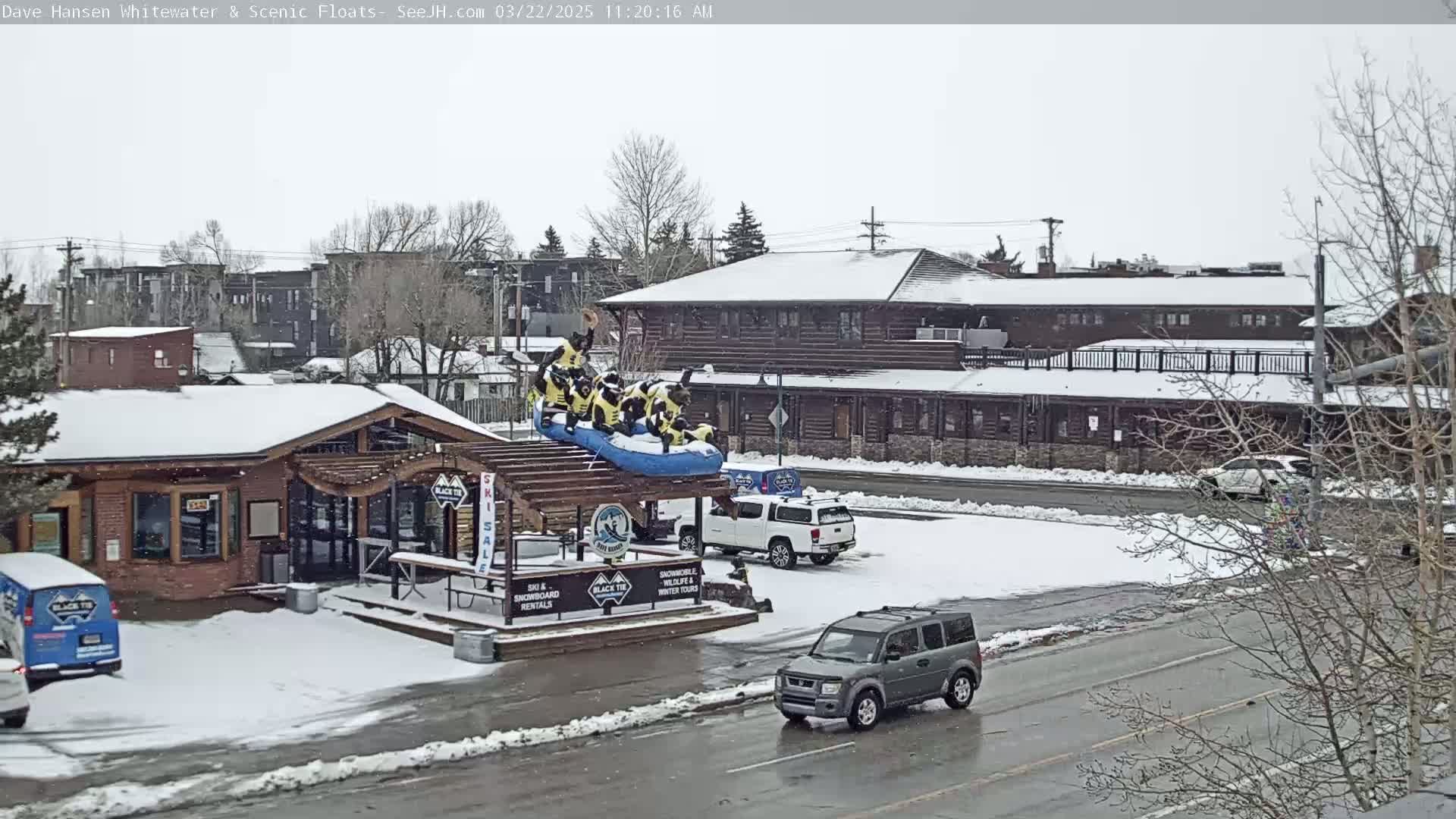 A snow-covered scene shows a building with a rafting display on the roof, a few cars in a parking lot, and bare trees in the background during a light snowfall.