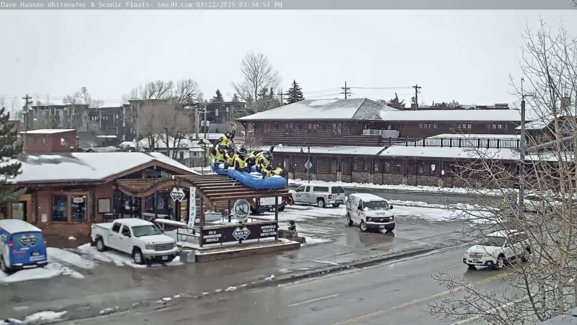 A snow-covered street scene shows a raft with people in it perched atop a building, several vehicles parked nearby, and light snow falling.