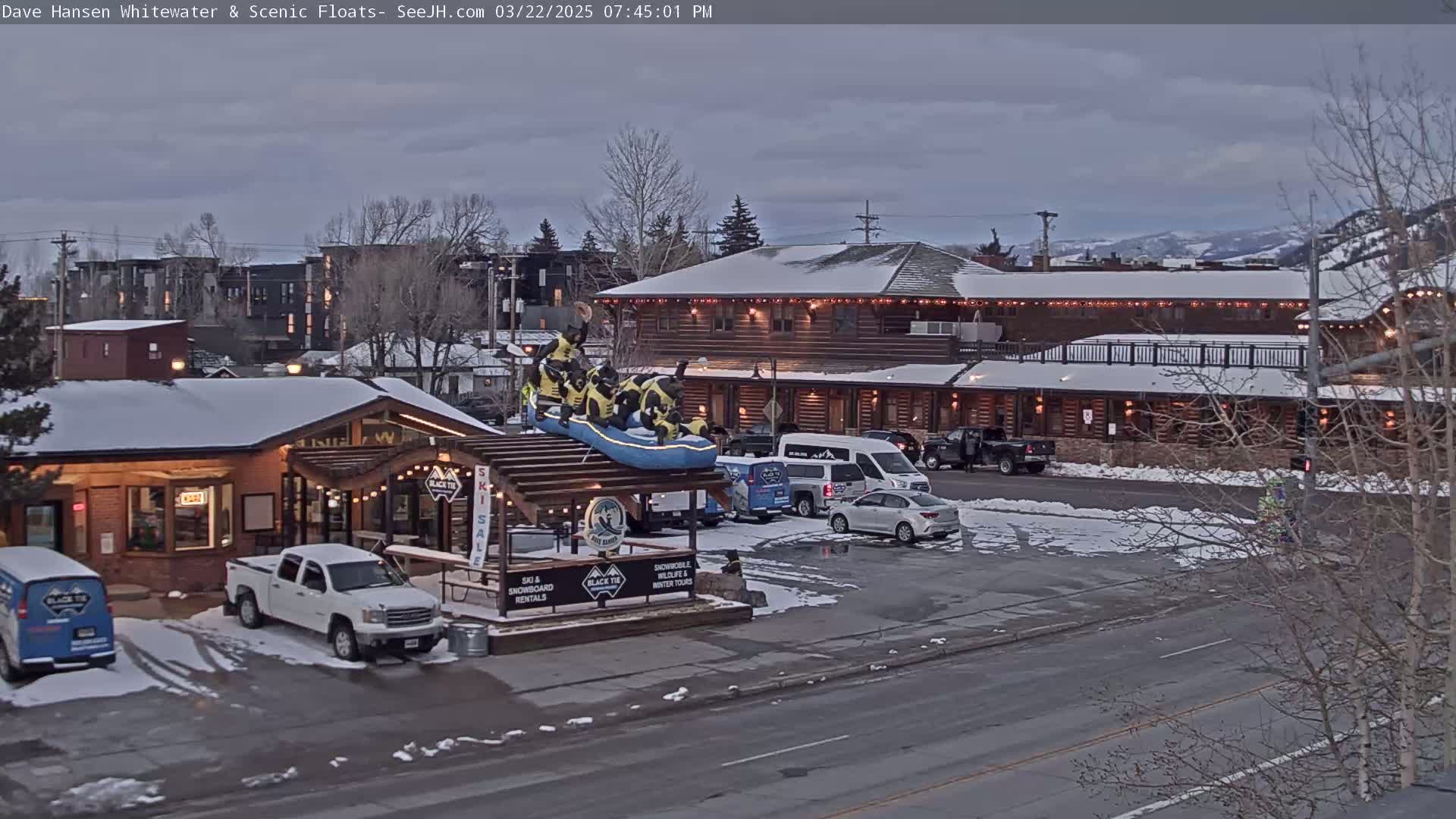 A snow-dusted parking lot in front of a log cabin-style building with several vehicles parked, a raft replica on a stand, and bare trees in the background under a cloudy sky.