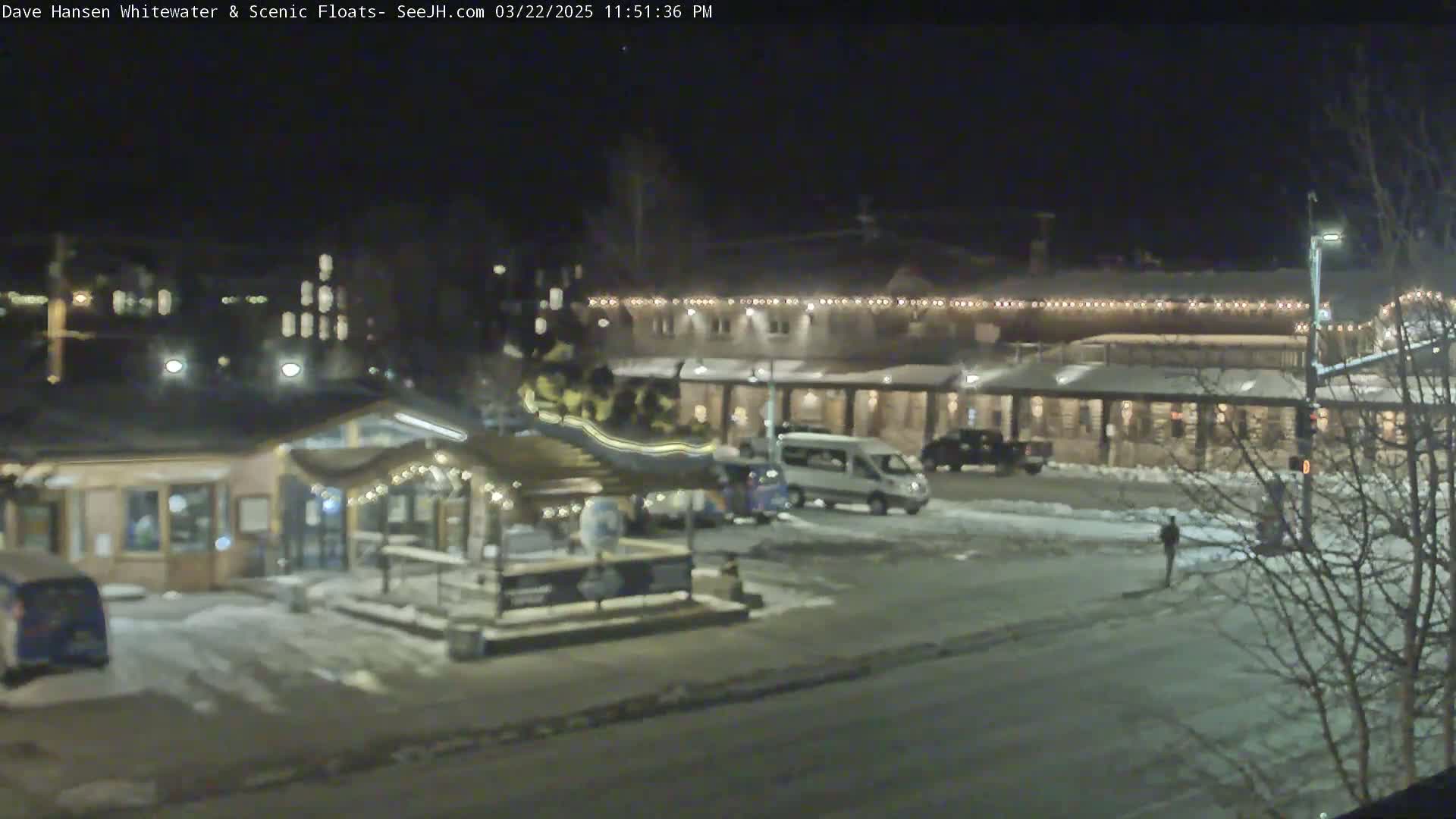A nighttime view of a snow-covered parking lot with several buildings, including a small kiosk and a larger building with lights strung along the roofline.