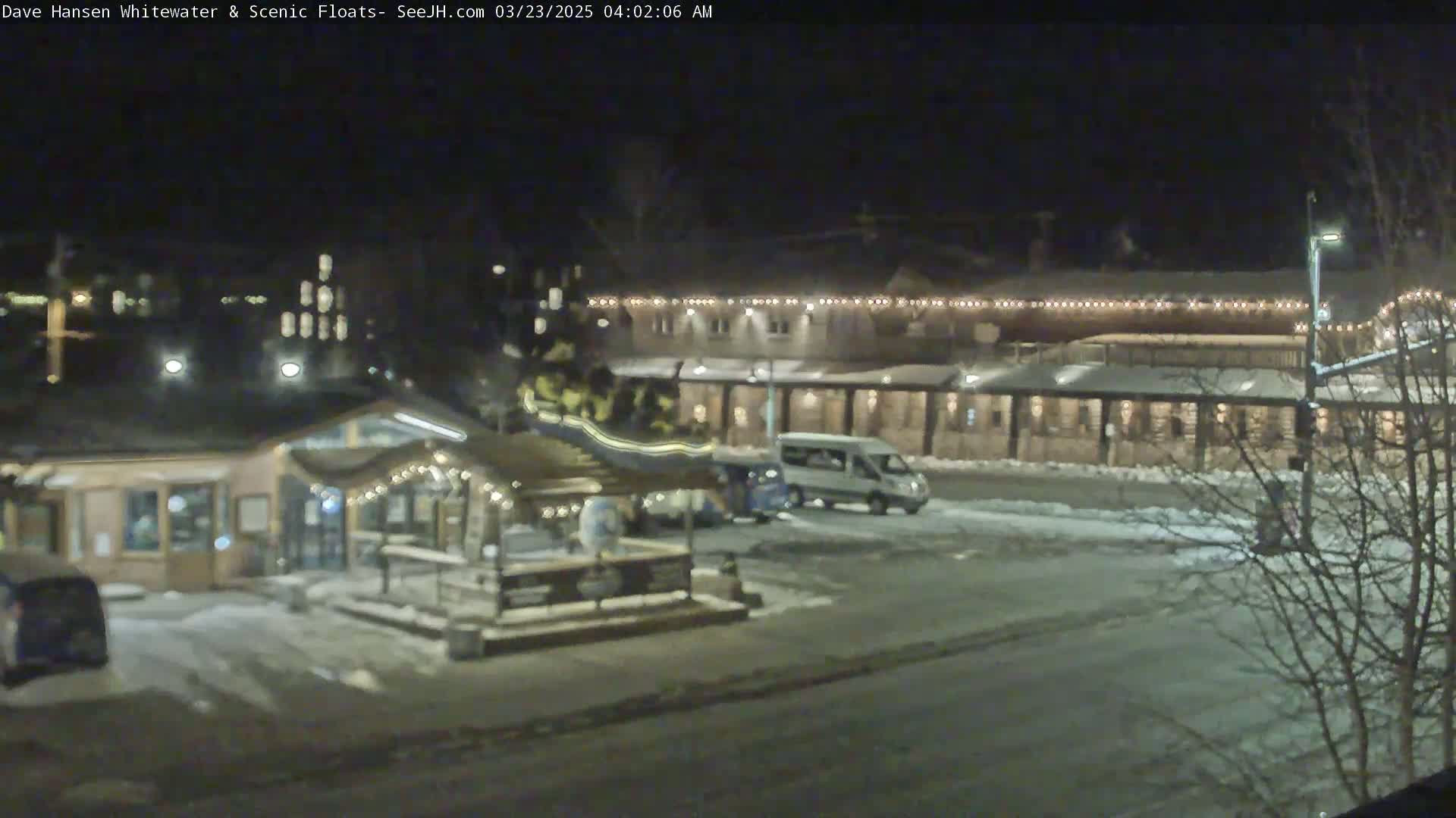 A nighttime view of a snow-covered parking lot with several buildings illuminated by lights, including a small structure with strings of lights and a larger building with a long row of lights along its roofline.