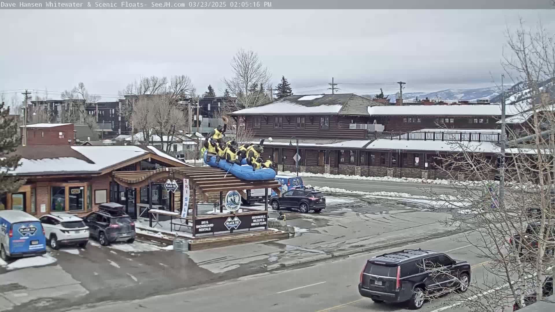 A group of people in yellow life jackets are on a raft atop a structure in a snowy parking lot, with cars parked nearby and buildings in the background under an overcast sky.