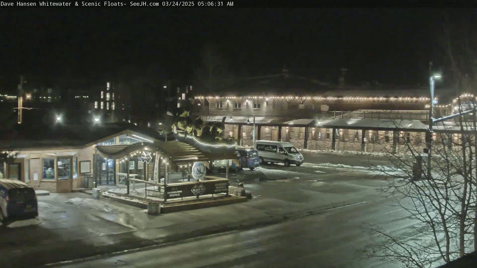 A nighttime, snowy scene shows a building with string lights, a parking lot with several vehicles, and a small structure with a sign.