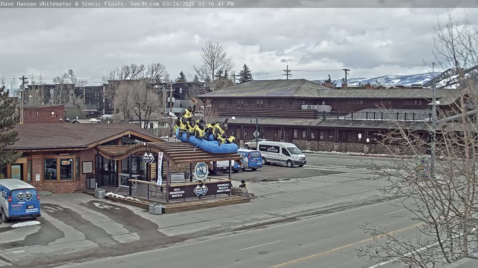 A partly cloudy day reveals a wooden building with a parking lot and a raft with bear figurines on top, situated near bare trees and snow-capped mountains in the distance.