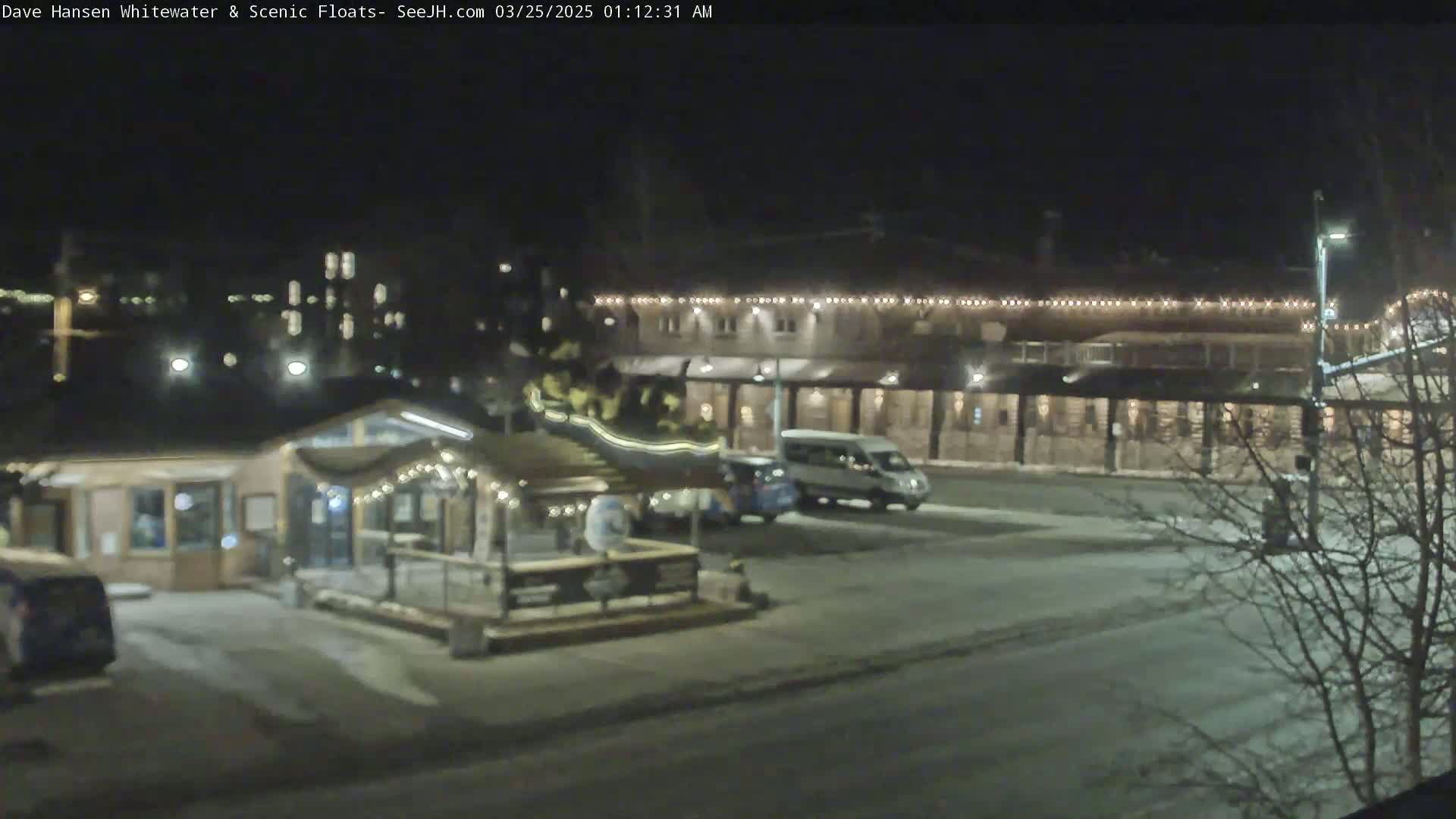 A nighttime view of a snow-covered parking lot with several vehicles, a small building decorated with lights, and a larger building with string lights across its roof in the background.