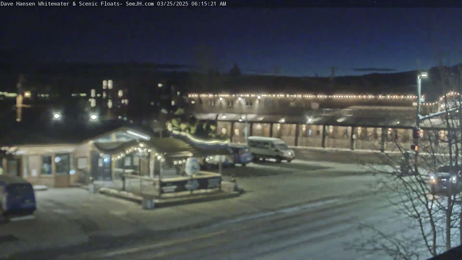 A nighttime view of a snow-covered town square with a building illuminated by string lights, several vehicles parked, and a body of water visible in the background.
