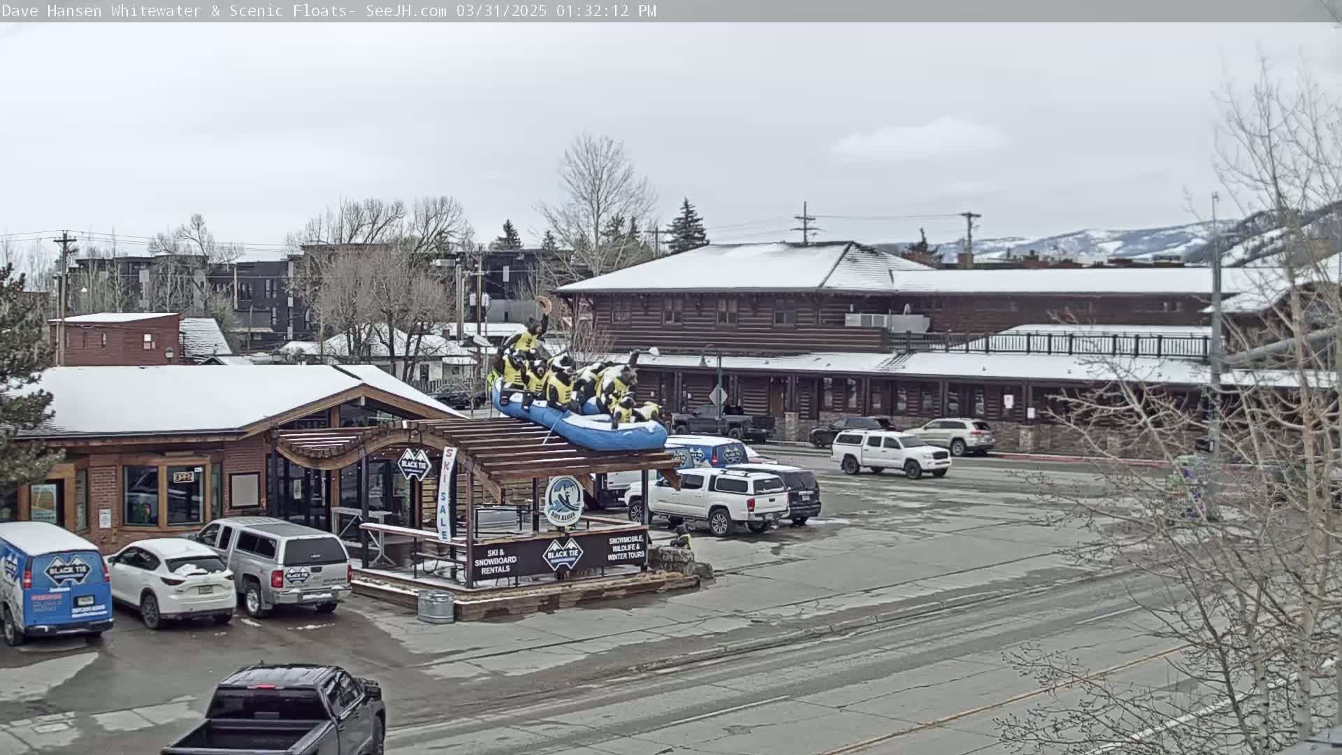 A snowy parking lot with several vehicles, a log cabin-style building, and a raft display in front of a rental shop.