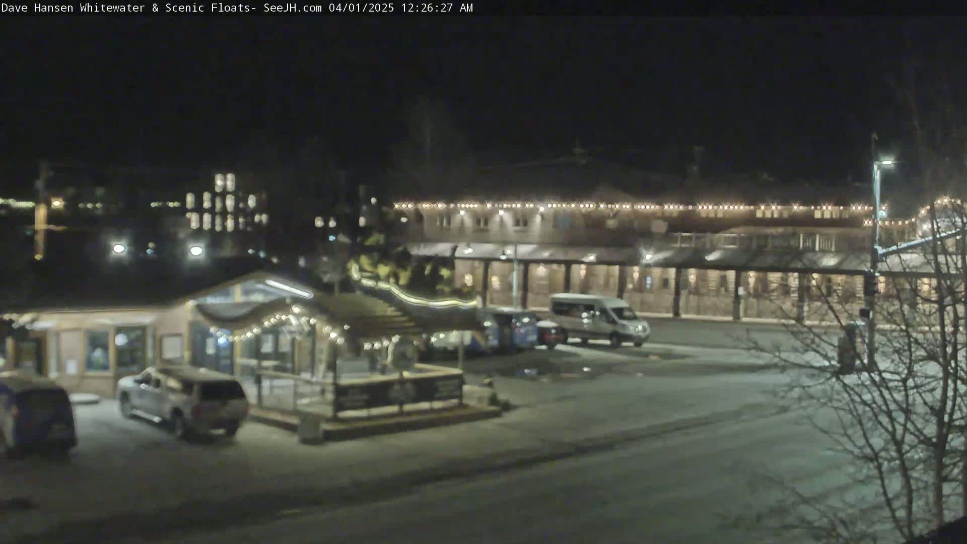 A nighttime scene showing a parking lot with several vehicles in front of a long, illuminated building and a smaller building with a covered patio area, all lightly dusted with snow.