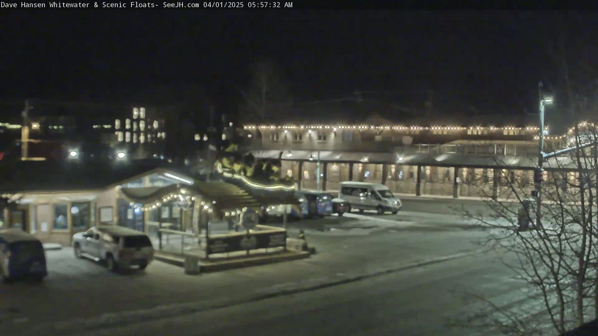 A nighttime scene of a snow-covered parking lot in front of a building with string lights, showing several vehicles parked and a smaller building with string lights in the foreground.