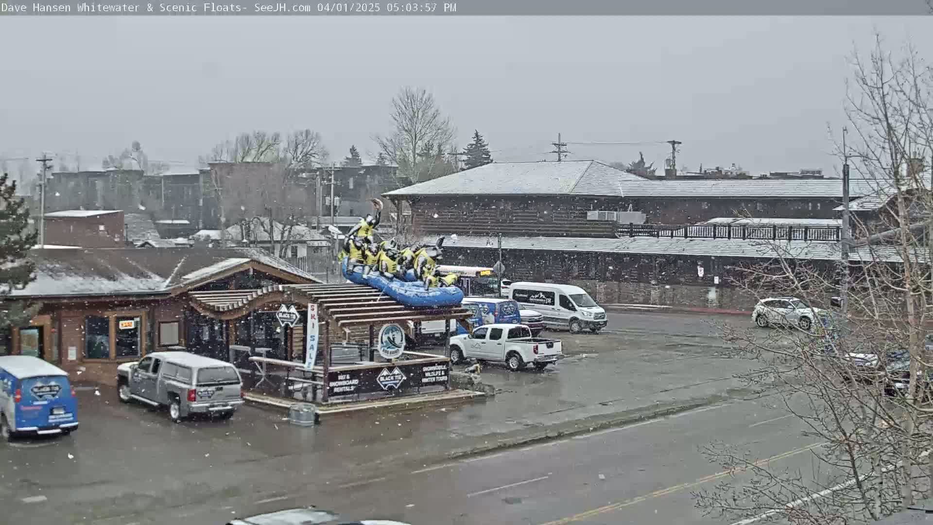 A snow-covered parking lot with a rafting display on top of a building, several vehicles, and a street scene in the background.