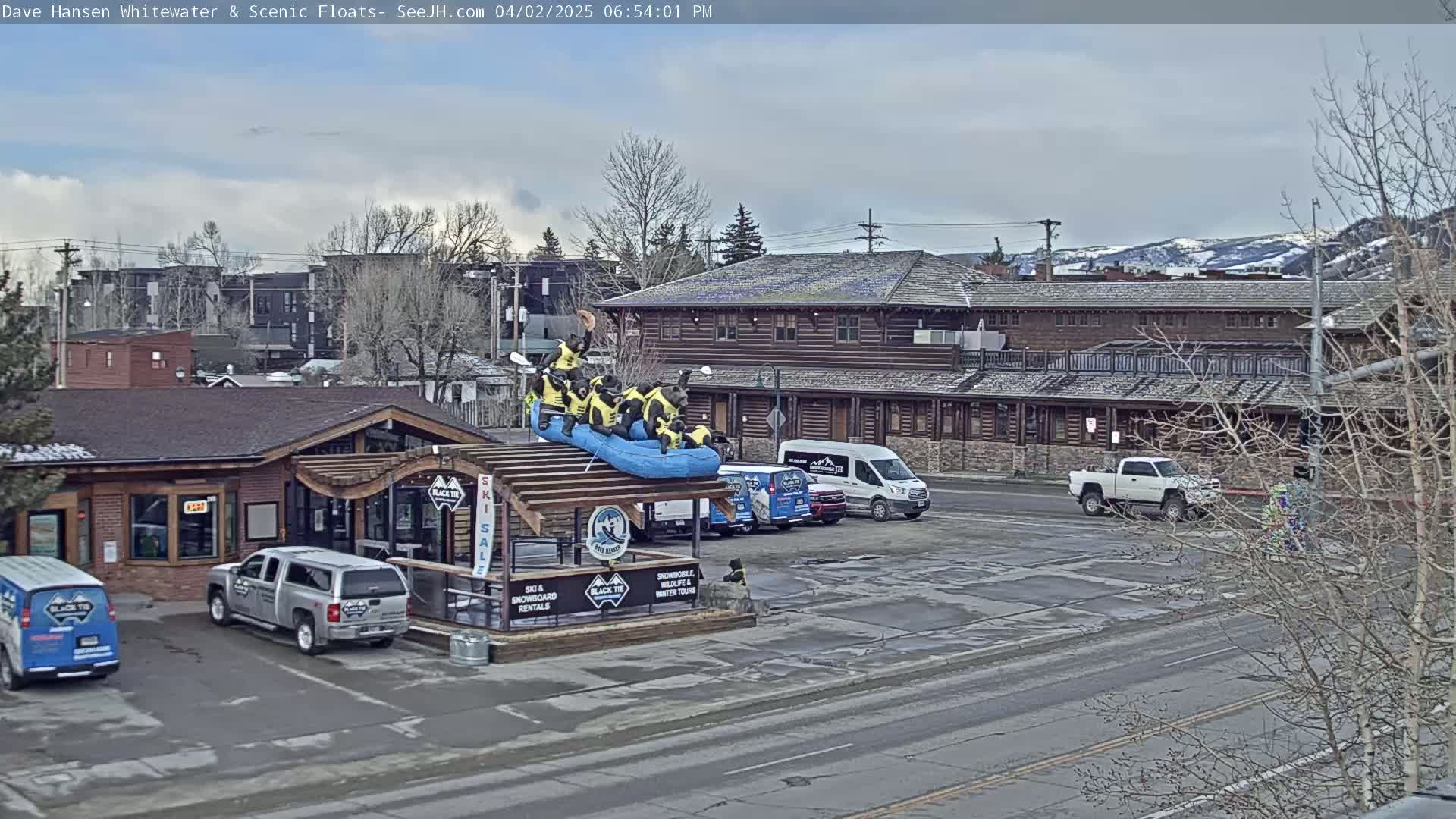 A partly cloudy day shows a storefront with a raft carrying bear figurines on the roof, several vehicles in a parking lot, and a building in the background.