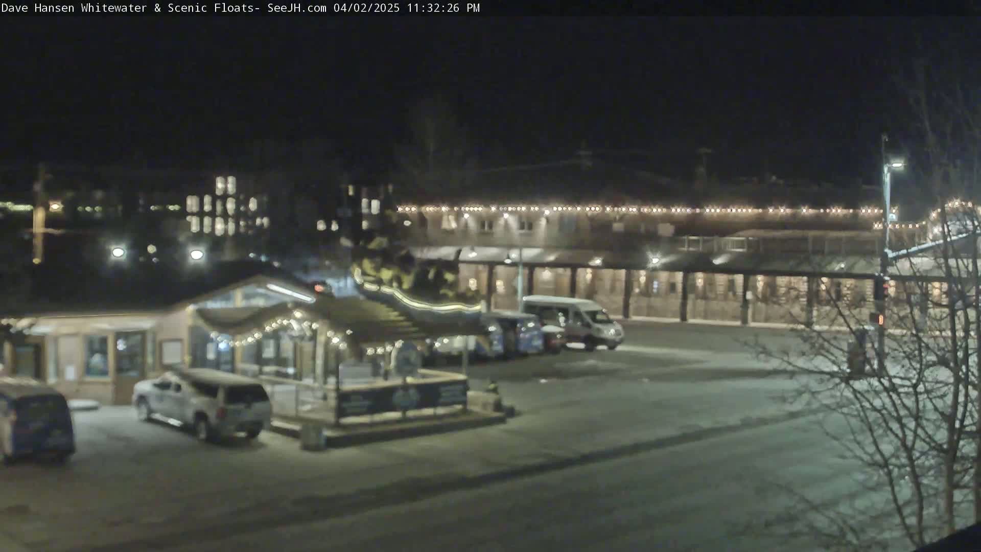 A nighttime view of a snow-covered parking lot with several vehicles parked near a building decorated with string lights.