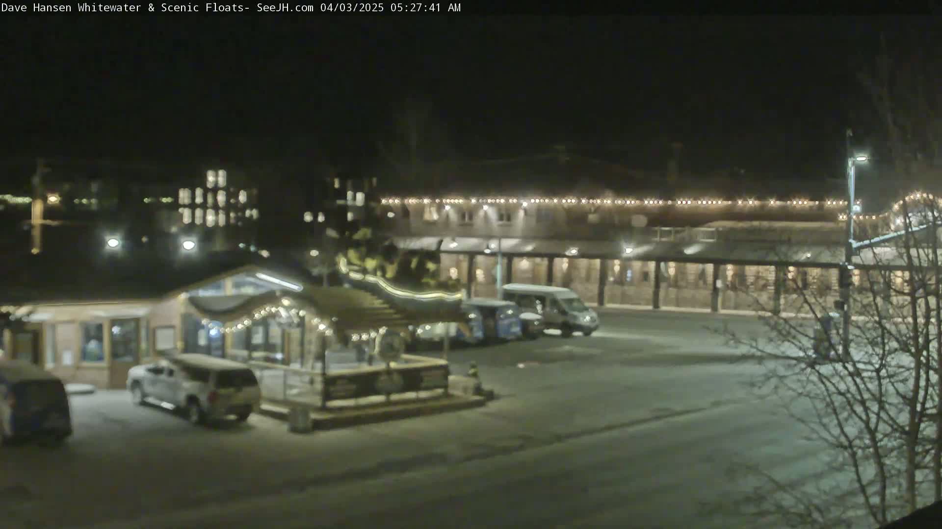 A nighttime view of a snow-covered parking lot with several vehicles parked near a building with lights strung along its roofline.