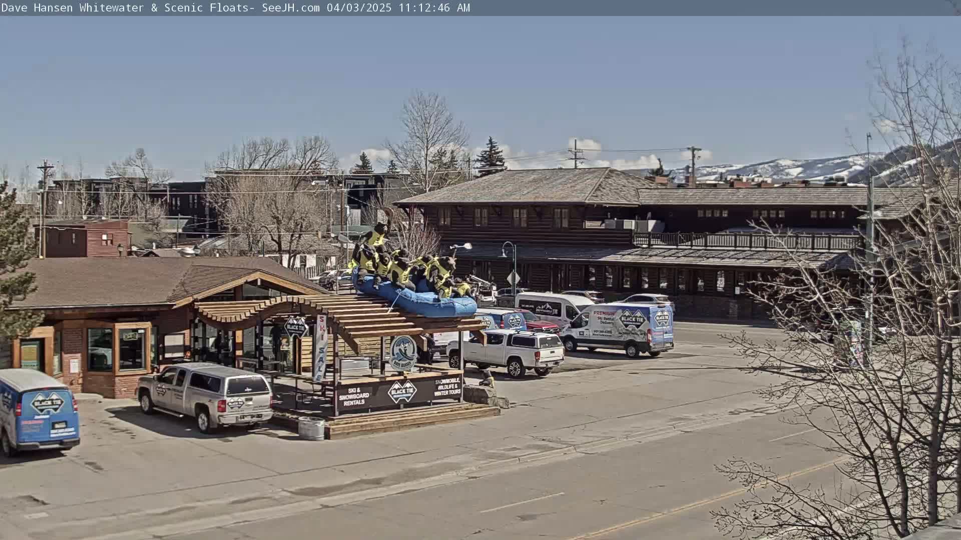 A sunny day shows a group of people on a raft atop a structure in a parking lot near a log building, with several vehicles nearby and snow-capped mountains in the distance.