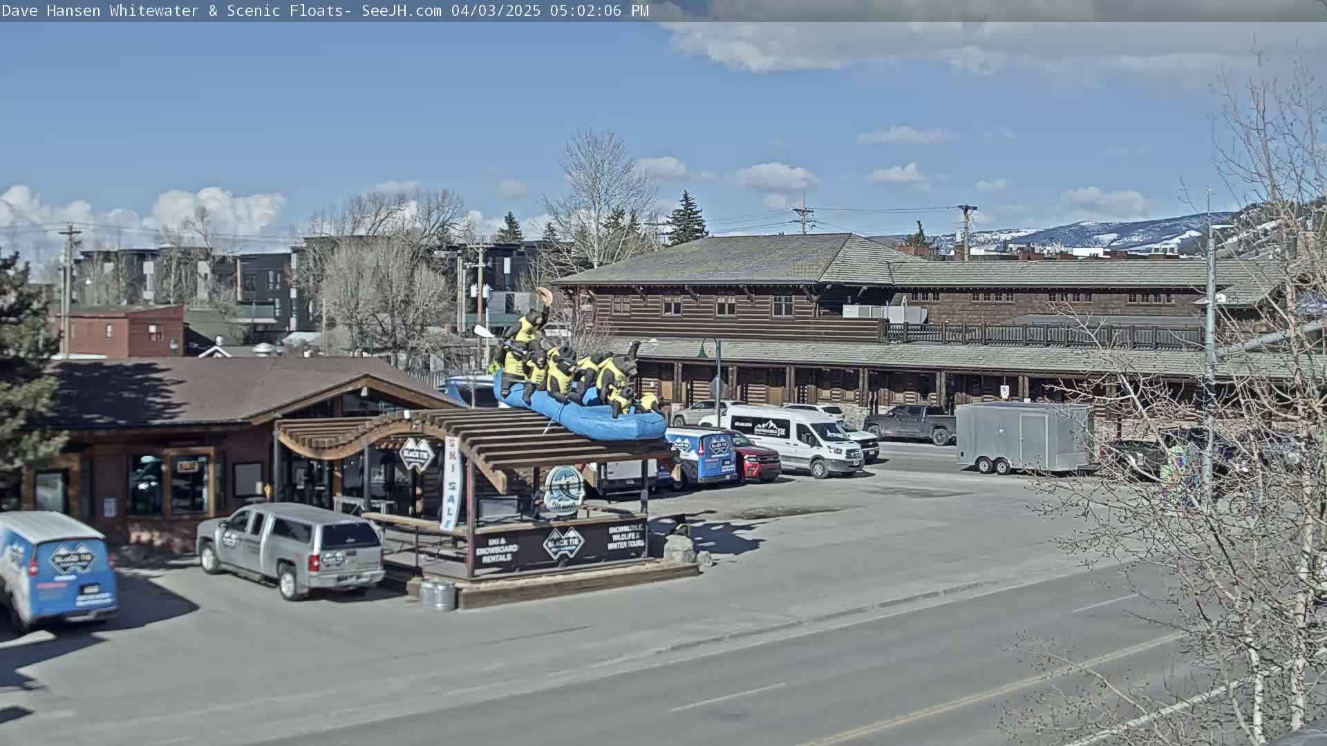 A sunny day shows a parking lot with several vehicles, a building, and a raft with bear figures on top of a structure in front of a building.
