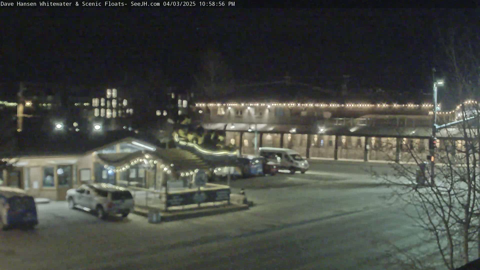 A nighttime scene shows a parking lot with several vehicles near a building decorated with string lights, with some snow or ice on the ground.
