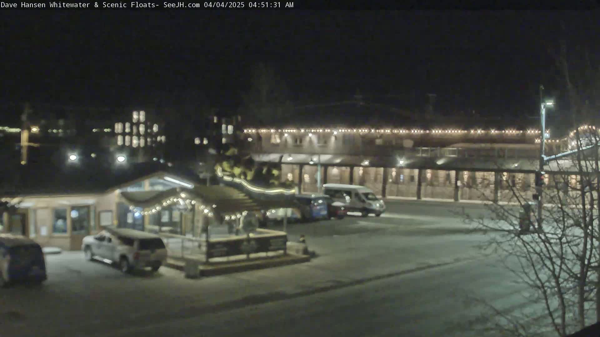 A snow-covered parking lot at night, with several vehicles parked near a building decorated with string lights.