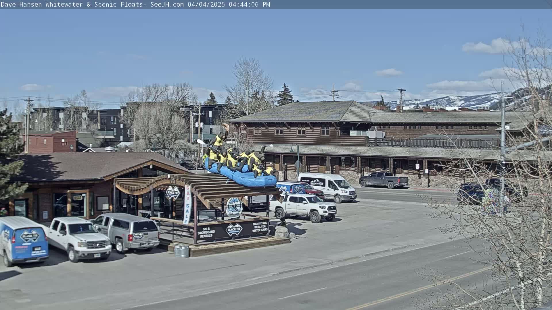 A sunny day reveals a parking lot with several vehicles, a building with a wooden exterior, and a display featuring a raft with bear figurines; snow-capped mountains are visible in the background.