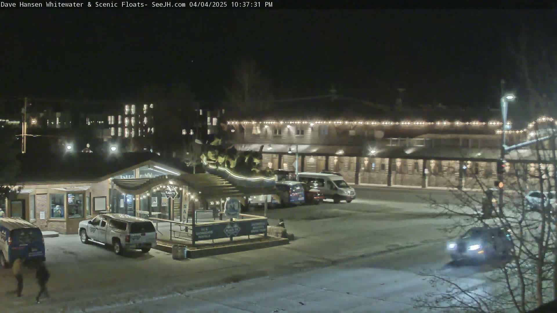 A nighttime snow-covered parking lot with several vehicles, a building with string lights, and a small structure with a ramp and signage.