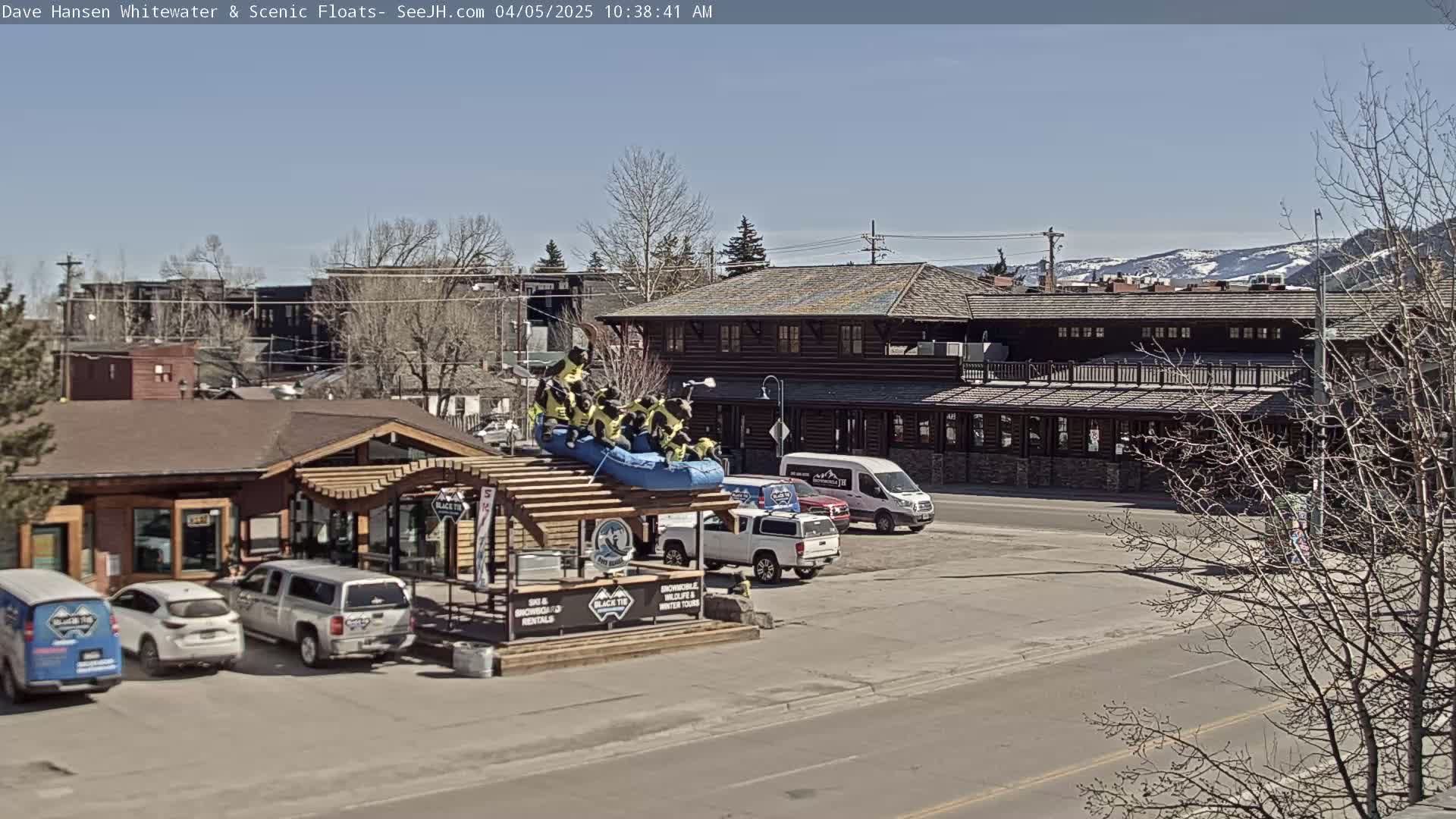 A sunny day reveals a street scene with parked cars, a log cabin-style building, and a rafting-themed display featuring a raft with figures atop a building.
