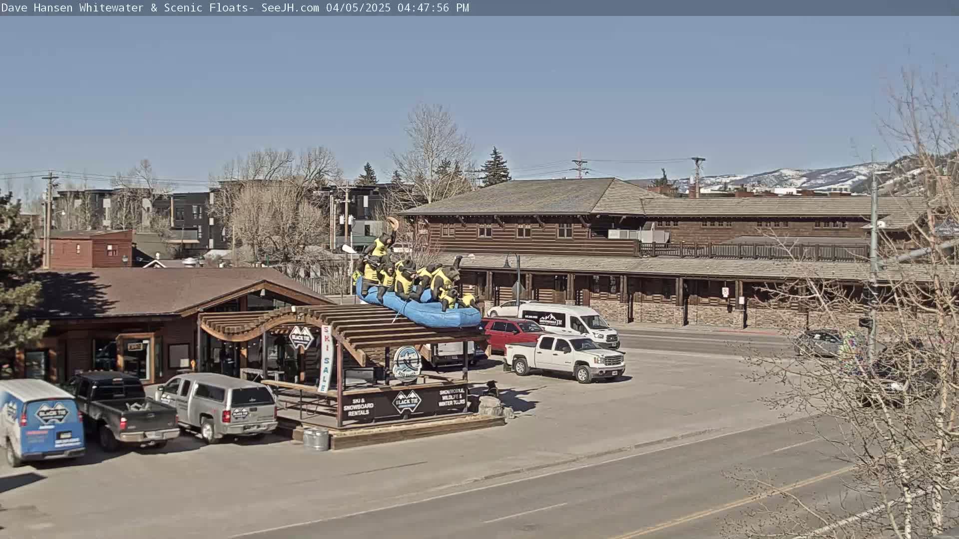 A blue raft with several figures in yellow sits atop a wooden structure in a parking lot on a sunny day, near several vehicles and a large, rustic-style building.