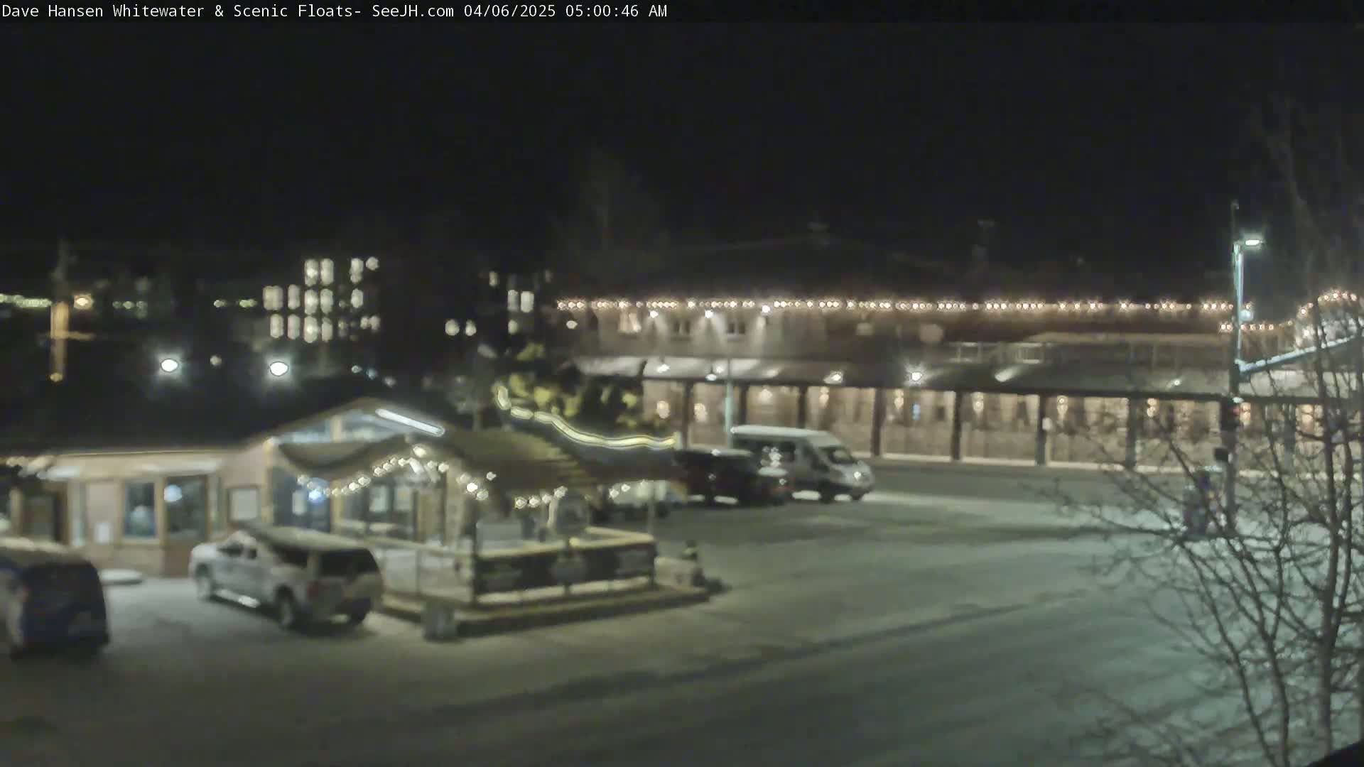 A nighttime view of a parking lot with several vehicles, a small building decorated with string lights, and a larger building with lights along its roofline, possibly in a snowy or icy area.