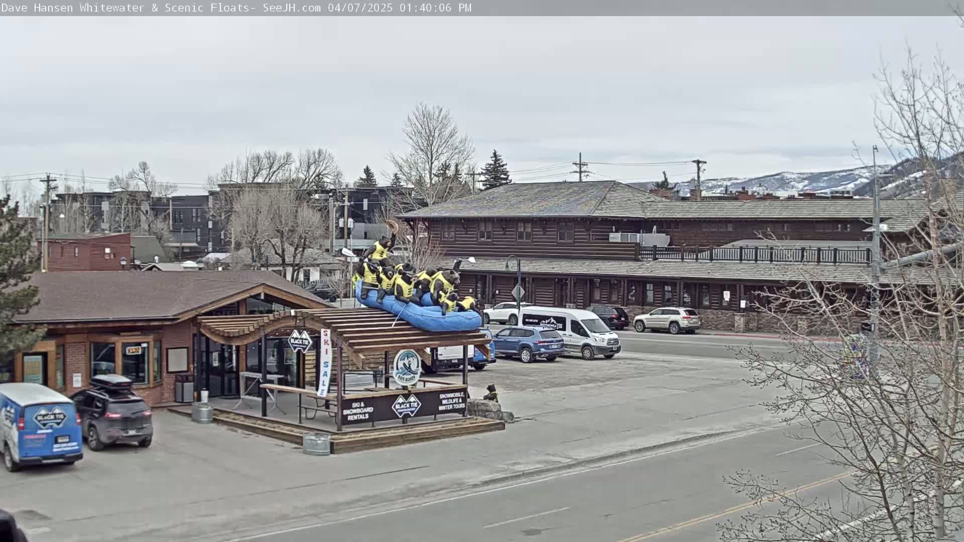 A mostly cloudy day reveals a riverside building with a raft sculpture of bears on top of a structure displaying outdoor recreation rentals, parked cars, and bare trees.