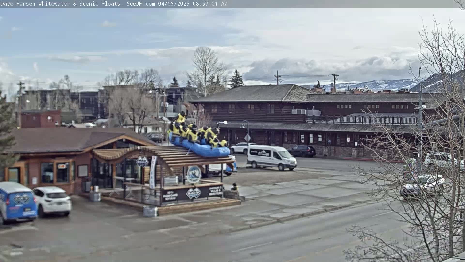 A partly cloudy day shows a river rafting company storefront with a raft on the roof, vehicles in a parking lot, and a long, dark-brown building in the background.