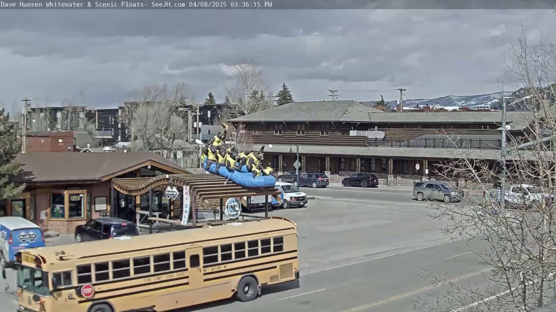Under a partly cloudy sky, a yellow school bus sits on a street in front of a wood-sided building where several people are on a raft atop a structure.