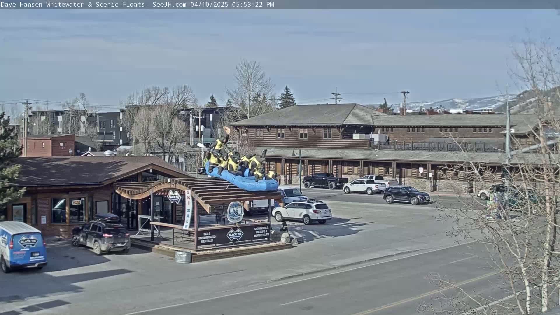 A blue raft with figures sits atop a wooden structure in a parking lot near a log building, under a sunny sky.