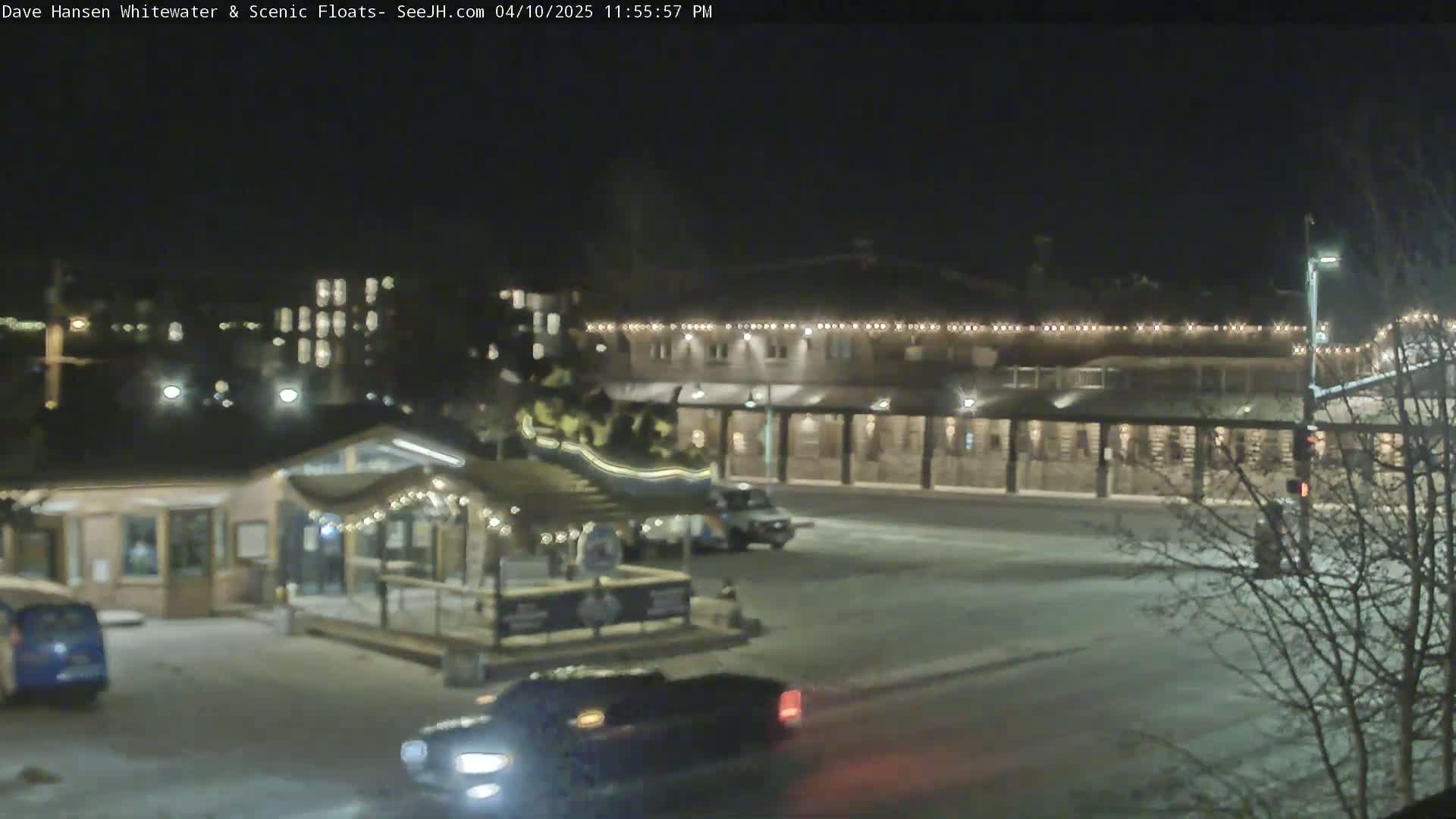 A nighttime, snow-covered parking lot with several cars and a building lit with string lights.