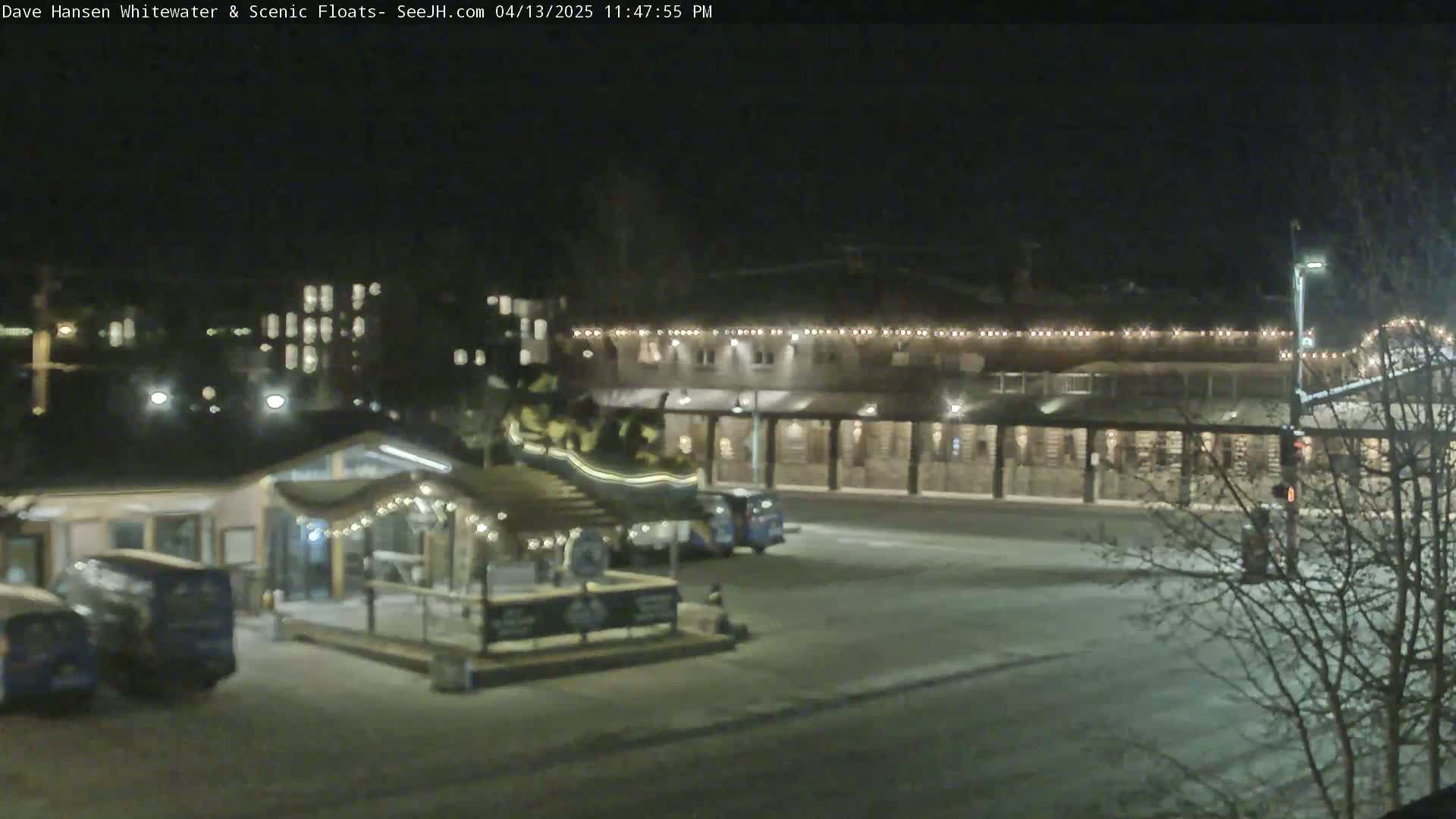 A nighttime view of a parking lot with several vehicles and buildings decorated with string lights, some snow on the ground.