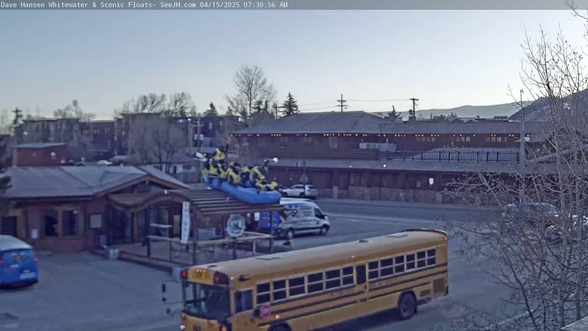 A yellow school bus is parked near a building with a group of people on a raft atop a structure, under a mostly clear, light-grey sky.