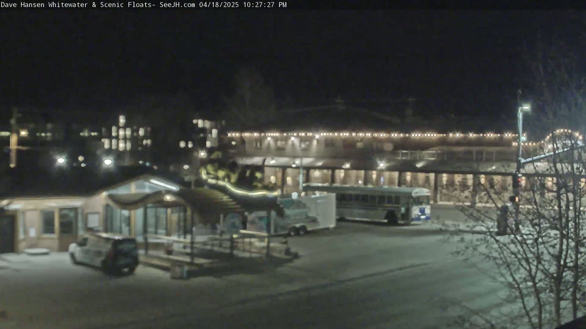 A nighttime view of a parking lot with a building illuminated by string lights, a small building, a bus, and several vehicles.
