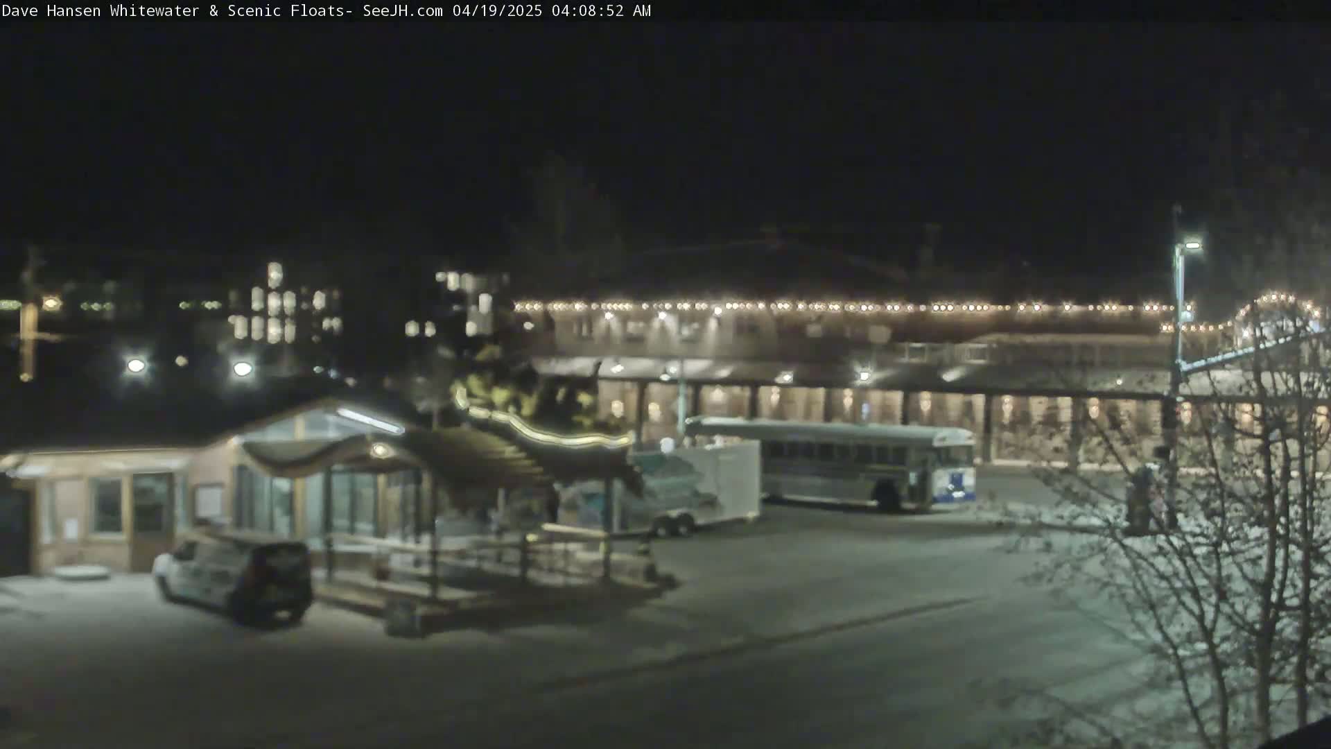 A nighttime view of a parking lot with a building lit by string lights, a small pavilion, a bus, and several vehicles.