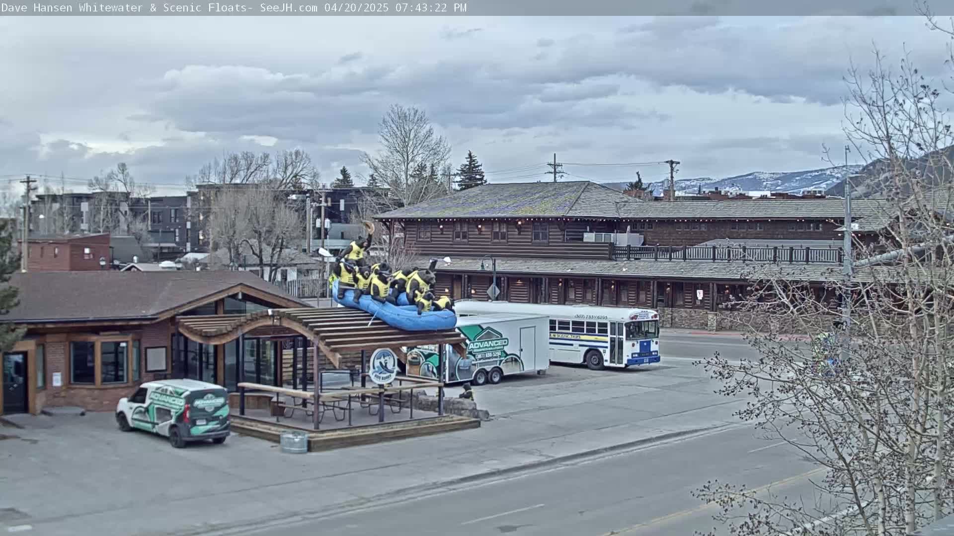 A partly cloudy day shows a group of people in a raft on a building's roof, a van, and a school bus parked in a lot near a log building.