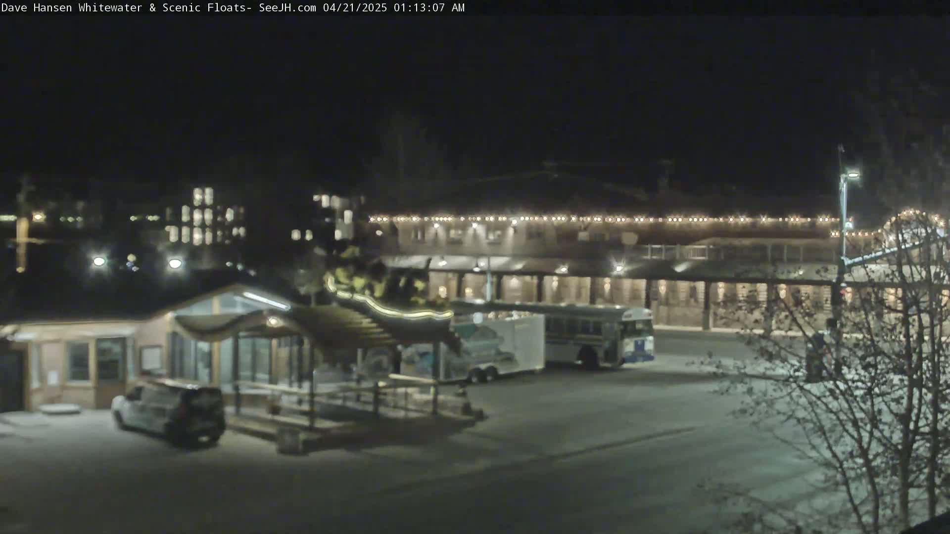 A nighttime view of a parking lot with a building featuring string lights, a small structure, several vehicles, and bare trees.