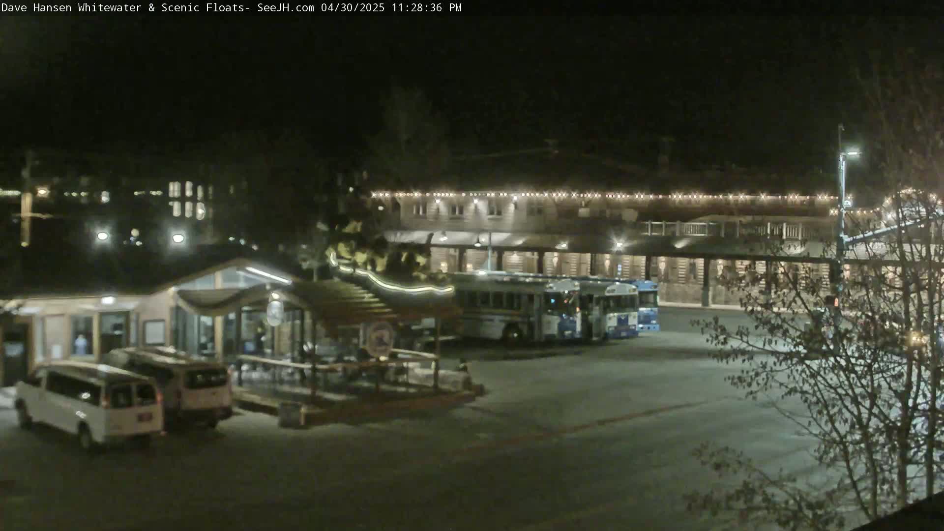 At night, a parking lot with two buses, two vans, and a small building with outdoor seating is illuminated under artificial light.