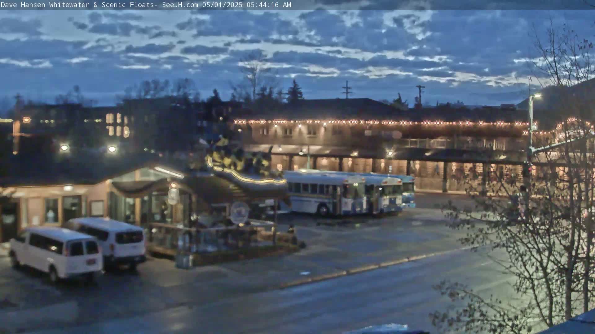 A pre-dawn scene shows a street with parked vans and a bus in front of a building lit with string lights under a partly cloudy sky.