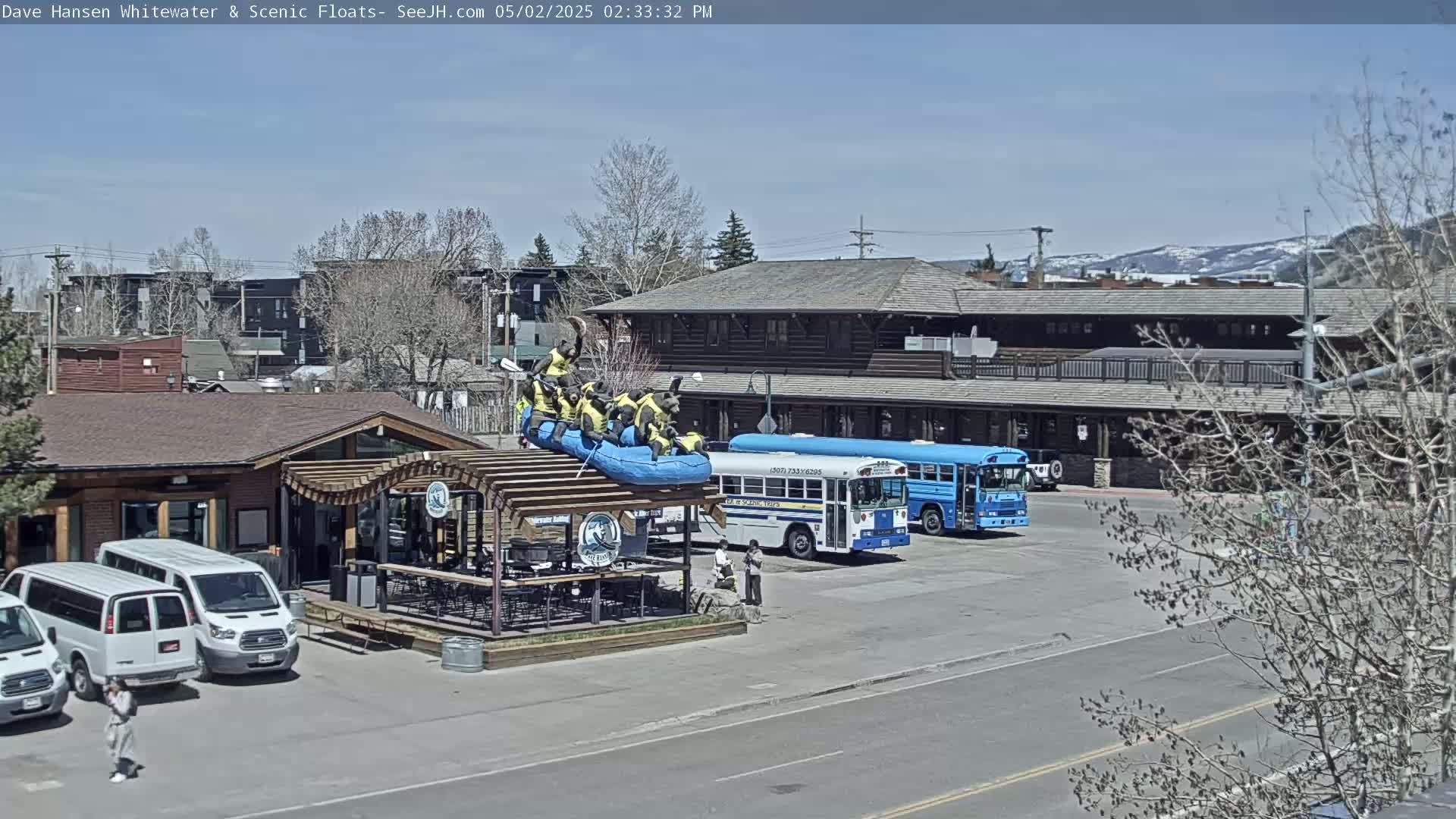 Under a sunny sky, a raft with bear figures sits atop a structure next to a building, with several vehicles including a blue school bus parked nearby.
