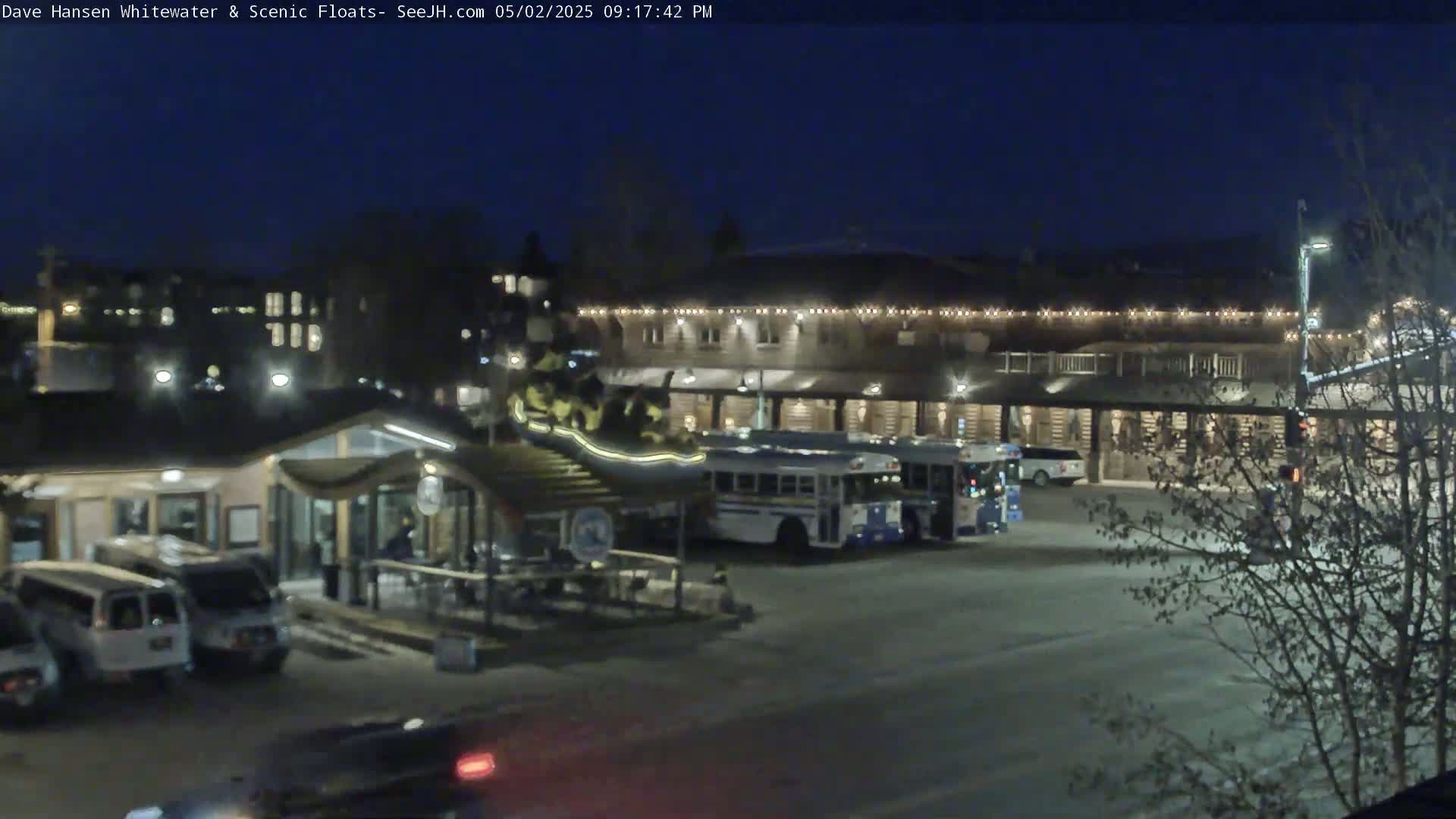 At night, a parking lot with several vehicles, including buses and SUVs, sits in front of a multi-story building illuminated with string lights.
