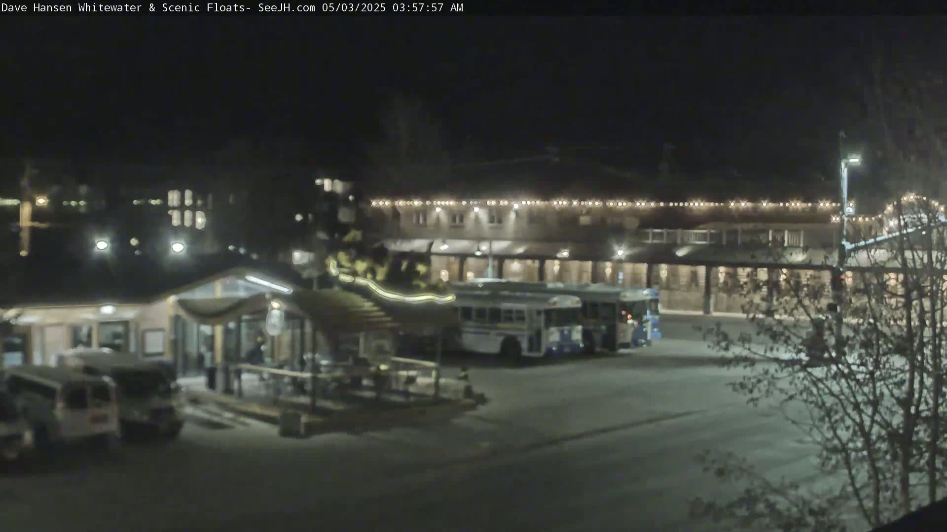 At night, a parking lot contains several vehicles, including two buses parked near a building with string lights outlining its roofline.