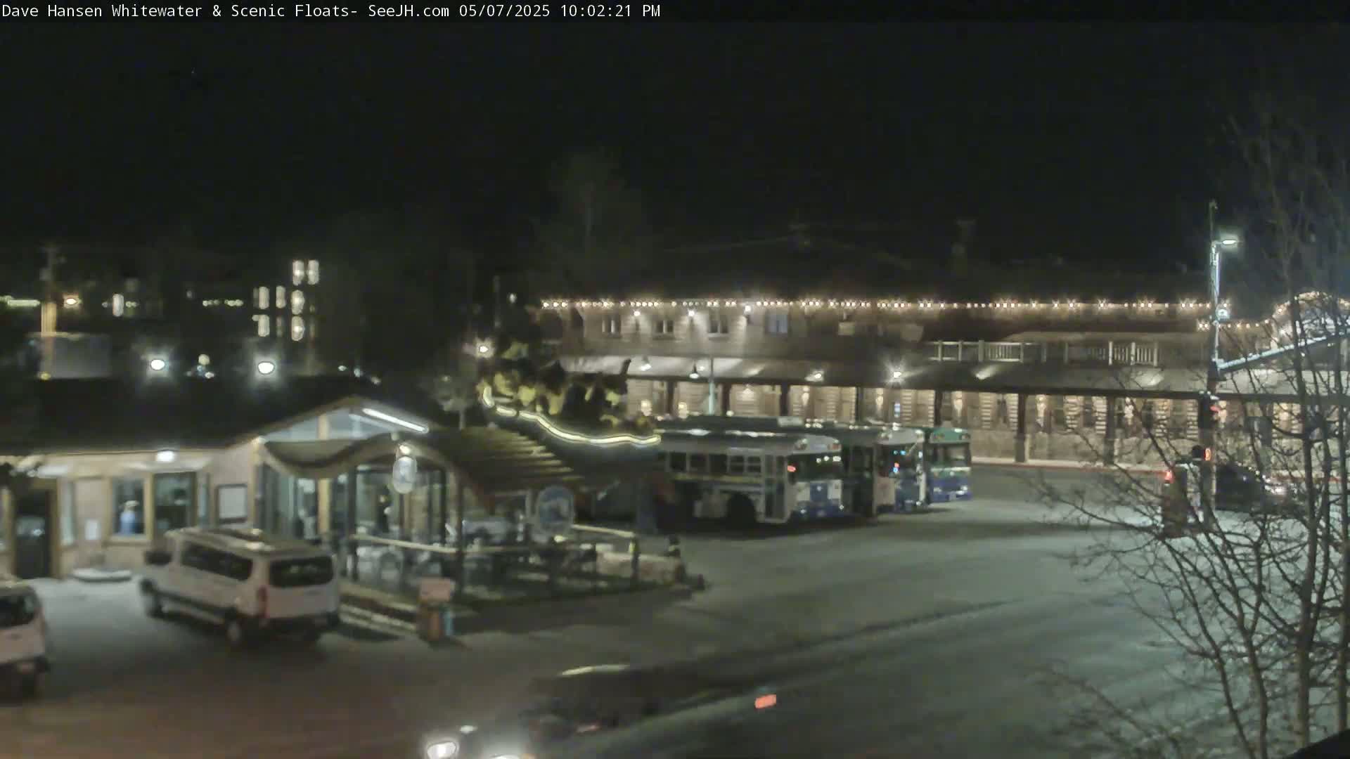 A nighttime view of a parking lot with several vehicles, including buses and vans, in front of a building decorated with string lights.