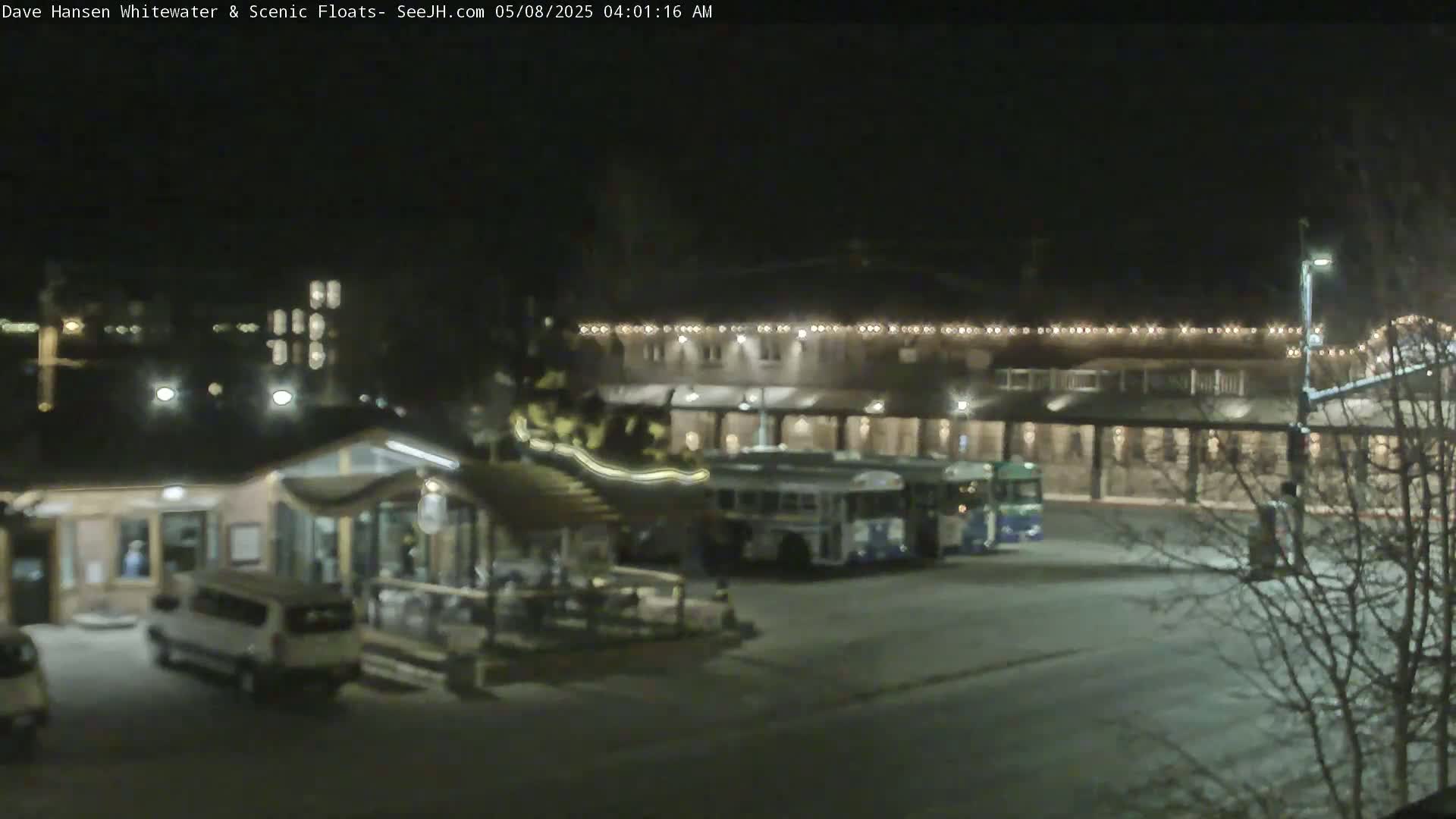 At night, several buses are parked in a lot next to a building with string lights, and a smaller building with a covered patio area is also present.