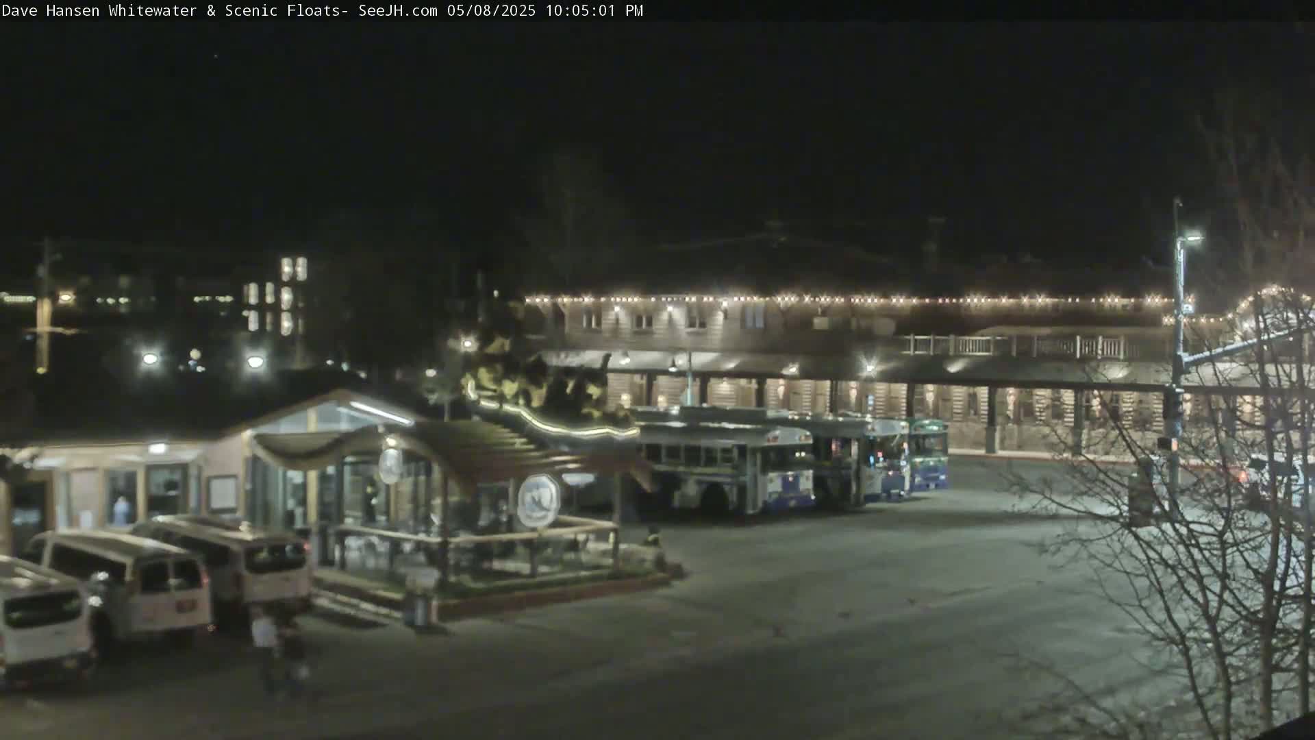 At night, a parking lot with several vans and two buses is in front of a building with string lights along its roofline, and bare trees are visible in the foreground.